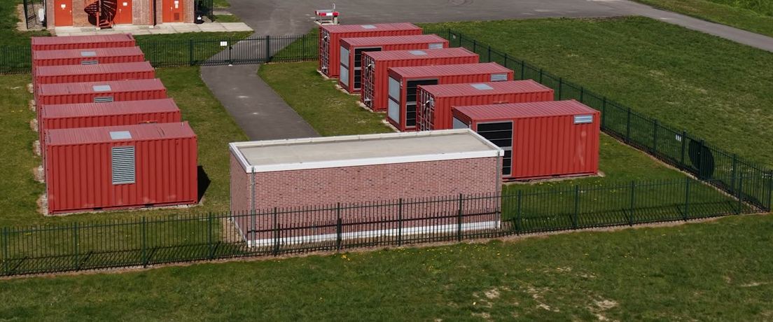 Container di accumulo rossi in fila su un prato recintato, con una cabina elettrica in mattoni davanti.